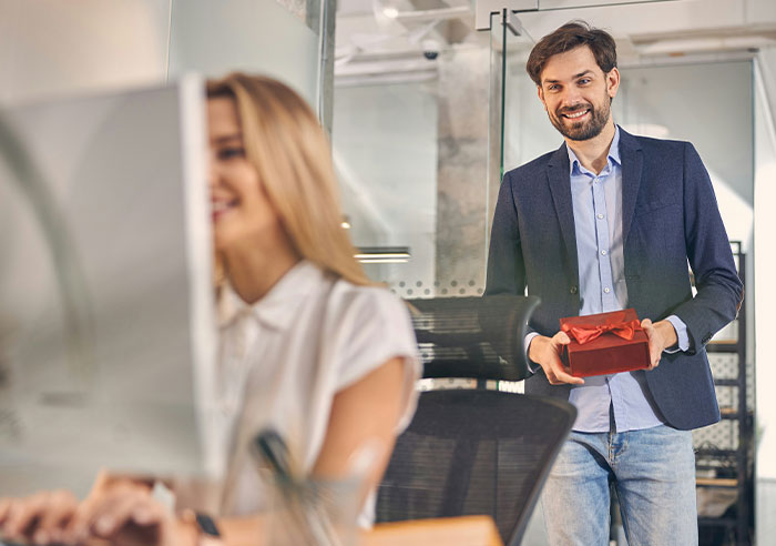 Man in a blazer holding a gift box, smiling at a woman working on a computer in a modern office setting.