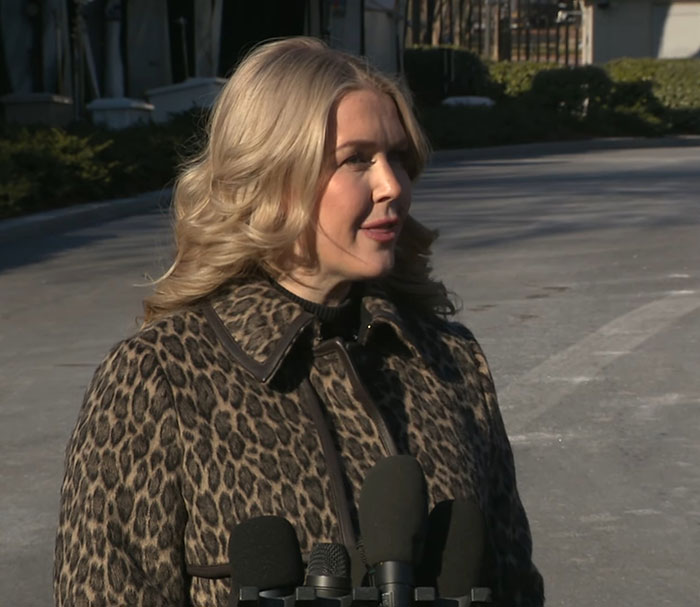 Karoline Leavitt speaking outdoors in a leopard print coat with microphones, VF photographer defends close-up amid mockery.