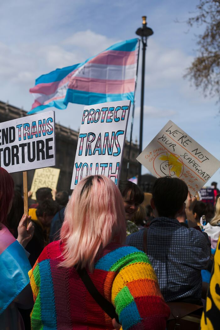 Protesters holding signs advocating for trans youth rights with a transgender pride flag waving in the background.