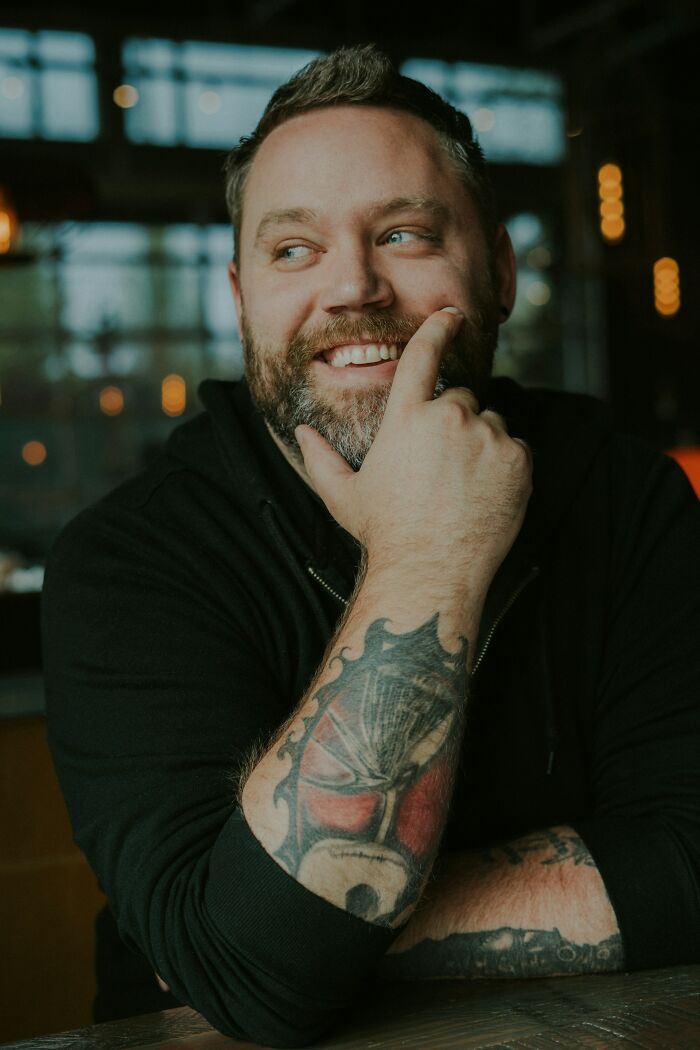 Bearded man with tattoos smiling thoughtfully, embodying the spirit of brilliantly sneaky acts of revenge.