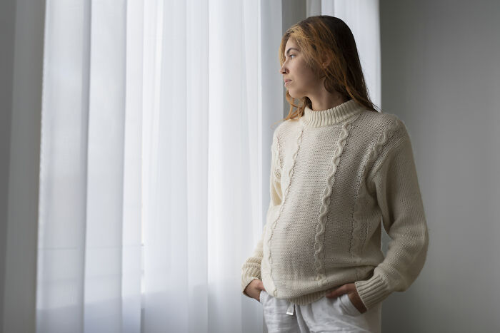 Young woman in a cream sweater looking pensively out the window, reflecting on a painful friendship breakup moment.
