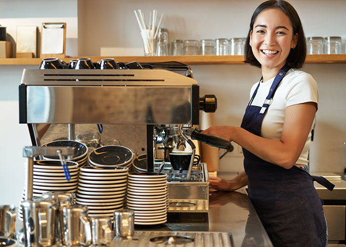 Coffee shop worker preparing espresso, smiling behind the counter with coffee machine and stacked cups in a busy cafe environment.