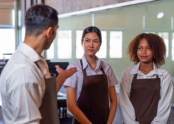 Coffee shop manager instructs workers on new rules while employees listen attentively during a team meeting.