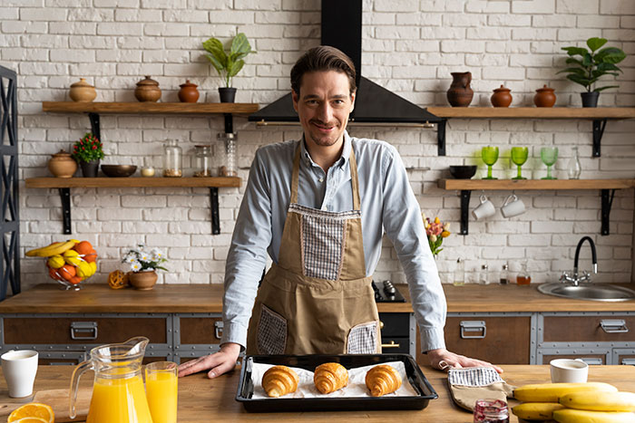 Man in apron presenting freshly baked pastries on tray in a kitchen, highlighting boyfriend's passion for pastries.