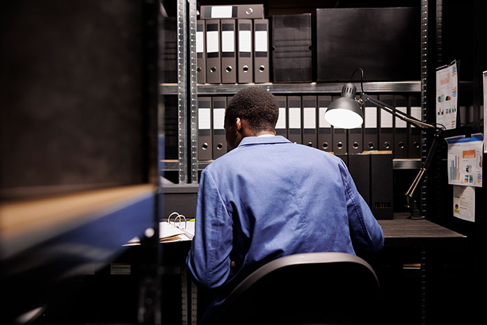Man in blue shirt sits at dark desk, surrounded by binders and documents, pondering wedding and private investigator findings.