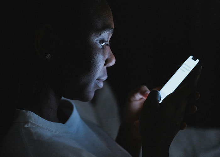 Woman in dark room focused on phone screen, illustrating phone dependence affecting family relationships.