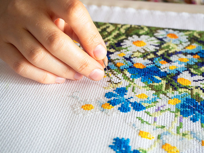 Hand stitching colorful floral cross-stitch embroidery on white fabric with focused detail and precision.