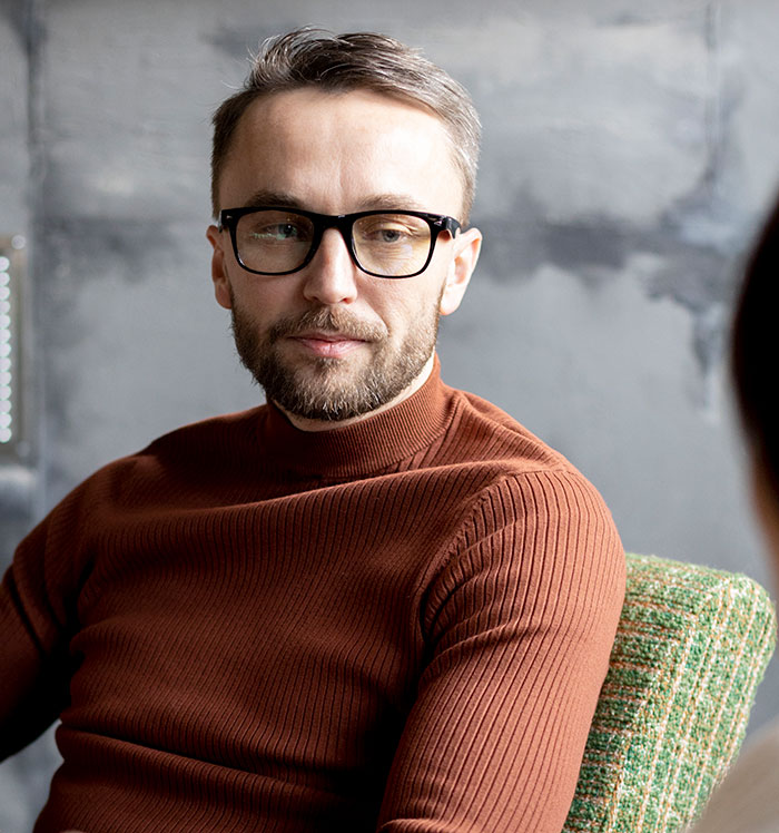 Man with glasses wearing a brown sweater mansplaining confidently to a woman in a casual indoor setting.
