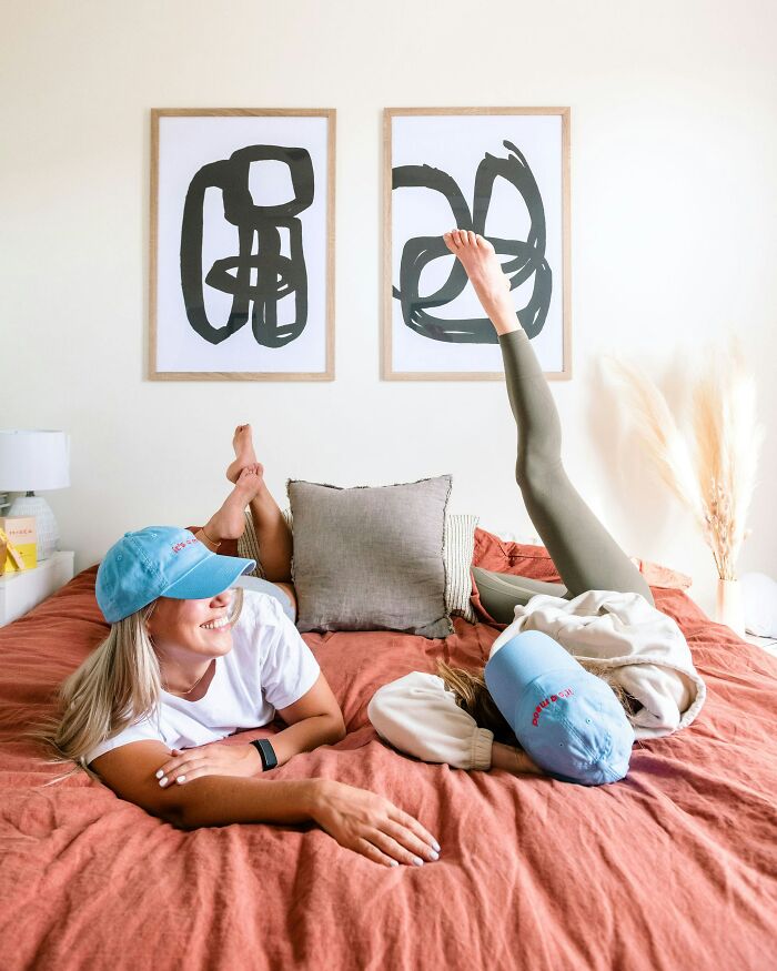 Two women wearing blue caps relaxing on a bed, highlighting the theme of distancing from a chaotic best friend.
