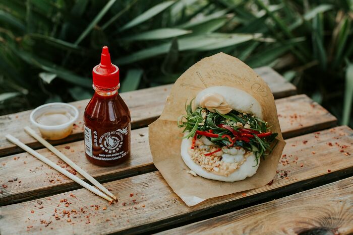 Bao bun with greens and sauce on a wooden table with chopsticks and a bottle, representing snobbish food presentation online.