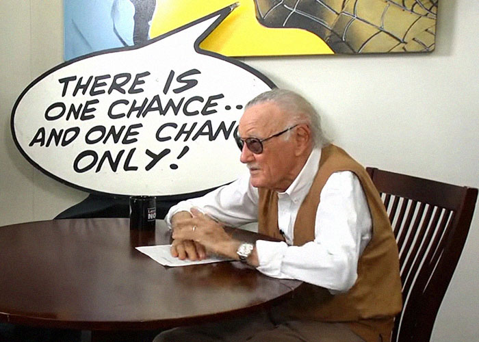 Elderly man wearing glasses and a brown vest seated at a table with a speech bubble discussing famous people they've met.