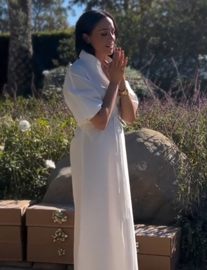 Meghan Markle in a white dress outdoors, appearing thoughtful, with rumors of Prince Harry split sparked by Christmas message