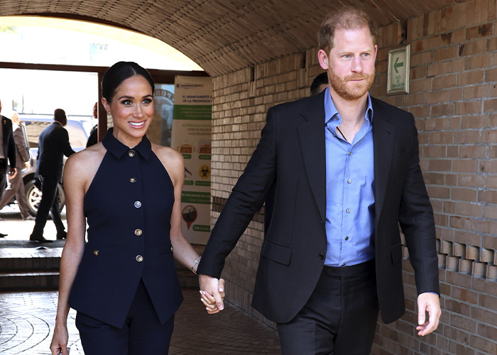Meghan Markle and Prince Harry holding hands, walking together in a public outdoor setting with smiles.