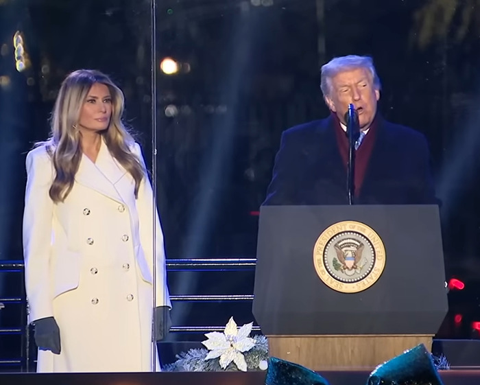 Melania and Donald Trump standing at a podium during a public event amid Christmas decorations and nighttime lighting