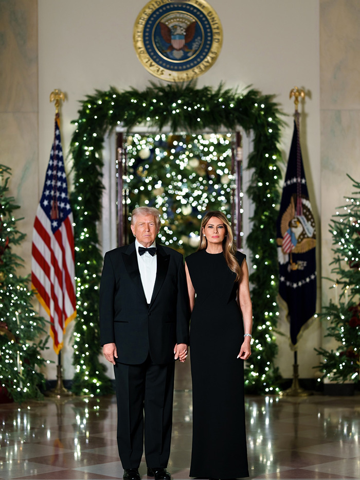 Donald and Melania Trump in formal attire posing for Christmas card with holiday decor and flags in background.