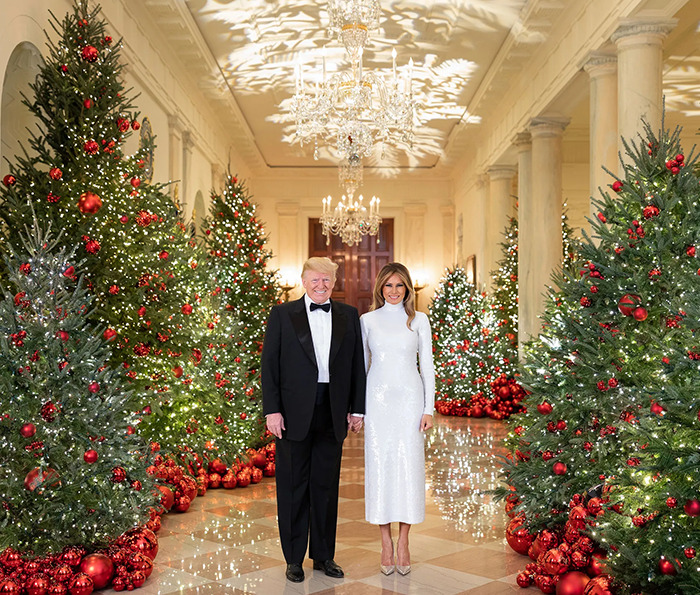 Melania and Donald Trump standing in a decorated hallway with Christmas trees in a holiday card scene.