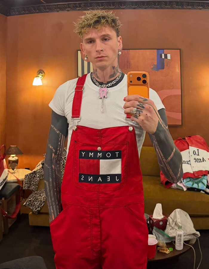 Machine Gun Kelly taking a mirror selfie wearing red overalls and a white shirt with tattooed arms visible. Machine Gun Kelly taking a mirror selfie wearing red overalls and a white shirt with tattooed arms visible.