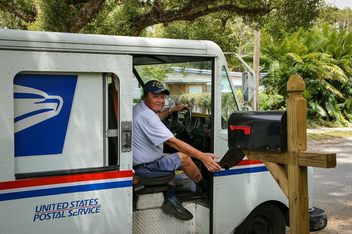 Postal worker in USPS vehicle delivering mail to residential mailbox on a sunny suburban street.