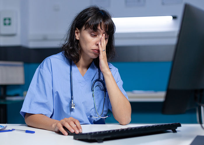 Tired woman in medical scrubs with stethoscope at computer, reflecting frustration after reheating pizza leftovers.
