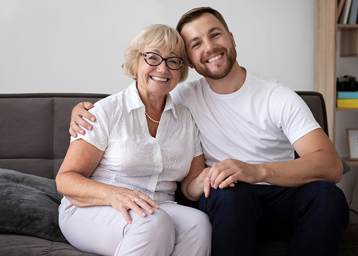 Smiling young man and older woman sitting close on sofa, representing woman reheats pizza leftovers for MIL during Xmas visit.