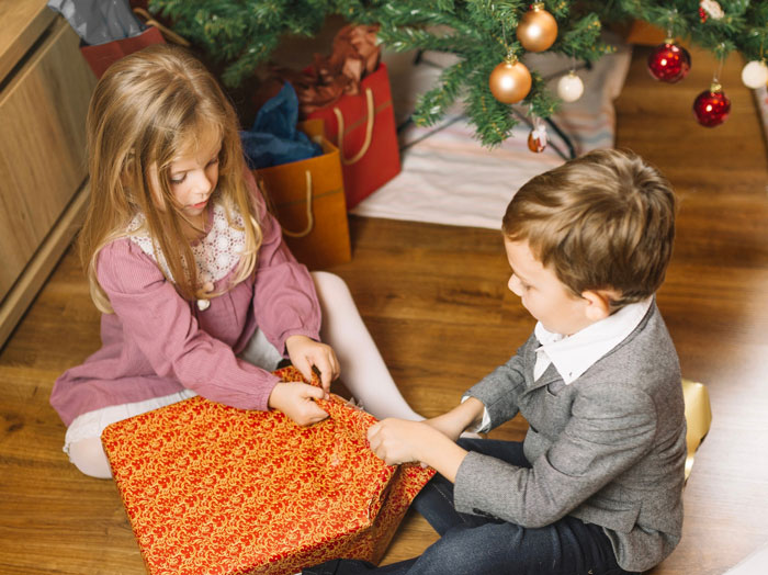 Two children sitting by a Christmas tree, unwrapping a large festive gift during holiday celebrations.