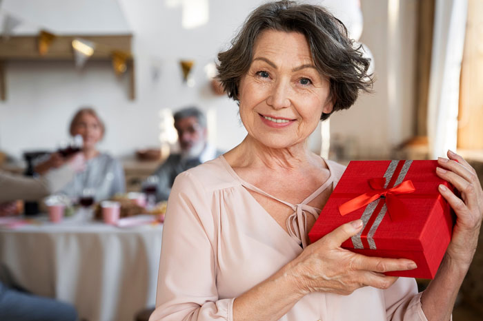 Older woman smiling and holding a red gift box during a family Christmas gathering at home.