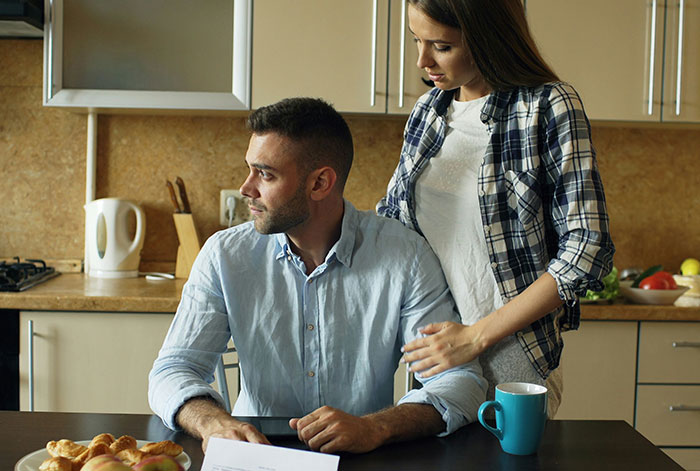 A woman confronts a man at the kitchen table, reflecting tension related to unhinged MIL losing her job and promotion news.