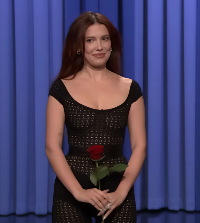 Actress in a sheer black outfit holding a red rose, highlighted in a studio setting with a blue curtain backdrop.