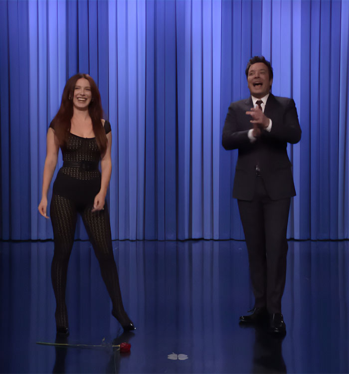 Actress in a sheer black outfit standing next to a host on a stage with blue curtains during a Fallon appearance.