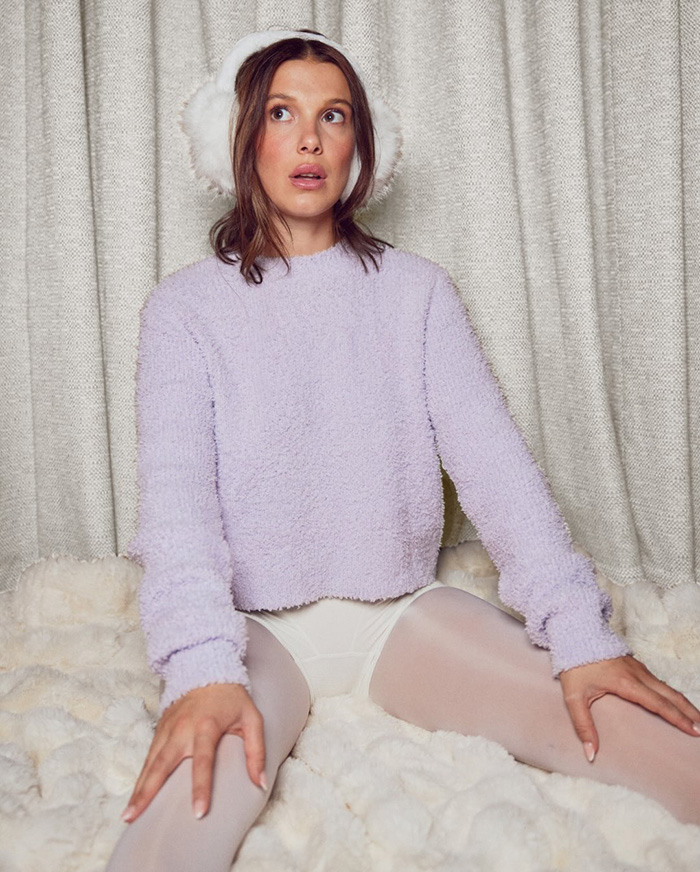 Young woman in lavender sweater and earmuffs sitting on faux fur with a snowy winter look sparking plastic surgery rumors.