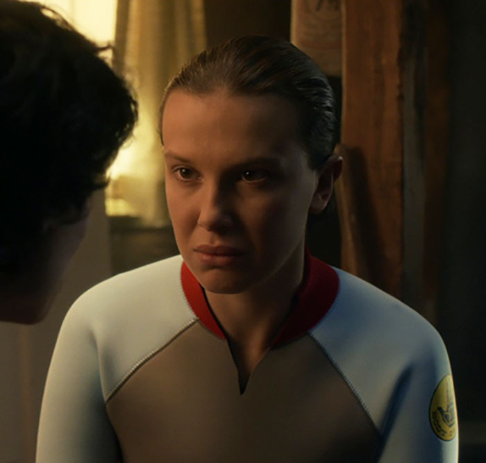 Millie Bobby Brown looking serious in a dimly lit room, reflecting on plastic surgery rumors and personal feelings.