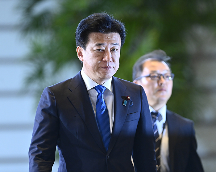 Japanese government official in a dark suit and blue tie walking with another man amid greenery background during a formal event