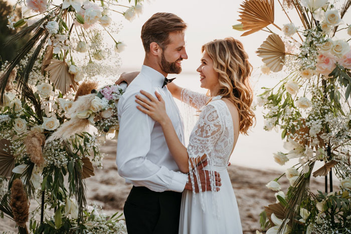 Bride and groom smiling at each other during outdoor wedding surrounded by floral decorations and greenery. Bride and groom smiling at each other during outdoor wedding surrounded by floral decorations and greenery.