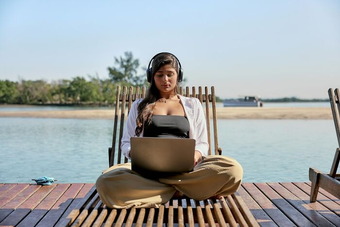Young digital nomad working on a laptop by the water, wearing headphones and sitting on a wooden deck outdoors.