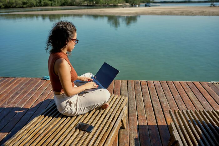 Woman working on laptop by the water, illustrating the digital nomad lifestyle away from typical Instagram posts.