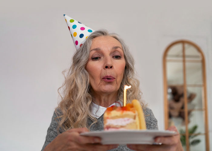 Older woman wearing a party hat blowing out a birthday candle, illustrating mom rages at son for forgetting birthday.
