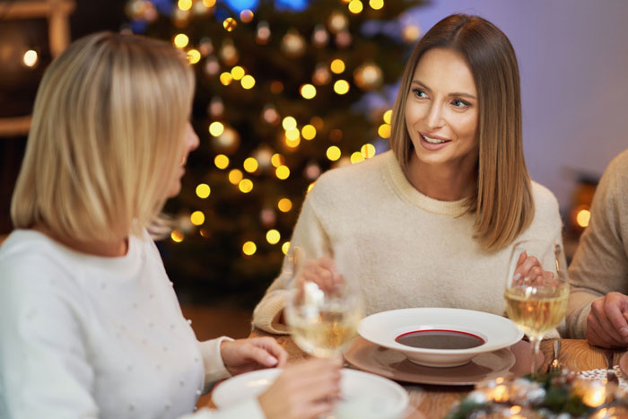 Two women enjoying Christmas dinner together, chatting and holding glasses of white wine in a cozy holiday setting.