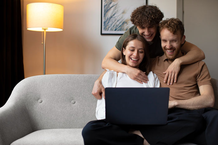 Young woman and two men smiling while using a laptop on a couch, illustrating family dynamics and relationship challenges.