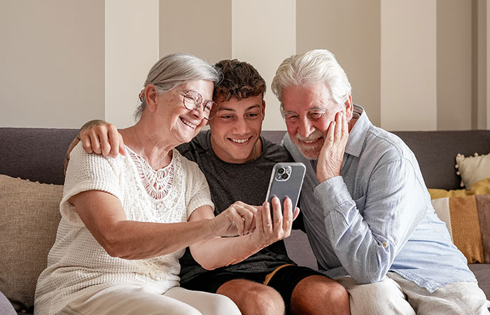 Exhausted teen smiling with grandparents on couch, all looking at smartphone. Exhausted teen smiling with grandparents on couch, all looking at smartphone.