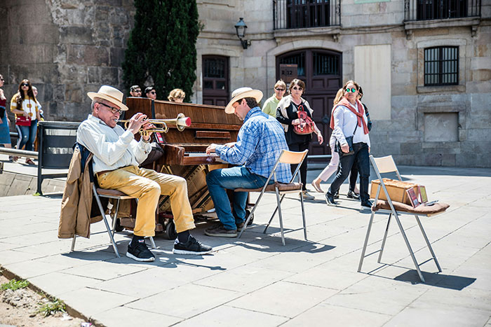 Street musicians from different countries play trumpet and piano in a public square while pedestrians walk by.