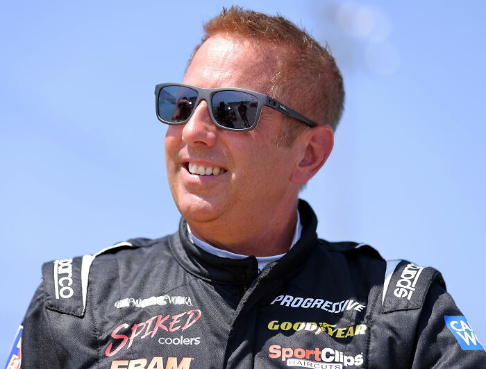 Race car driver Greg Biffle wearing sunglasses and a black racing suit outdoors on a sunny day