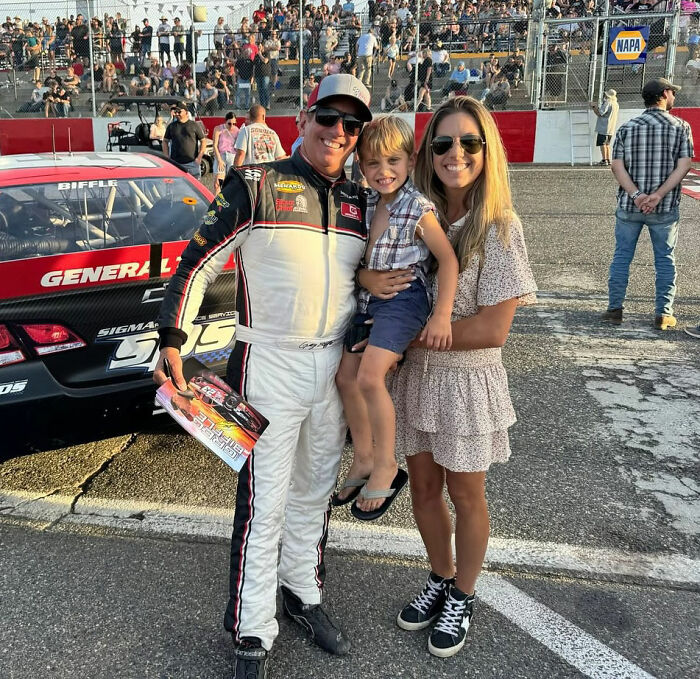 Greg Biffle with his wife and child at a race track, standing beside a race car with a large crowd in the background.