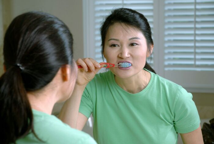 Woman brushing teeth in front of mirror, highlighting everyday things that are more dangerous for your health than you realize