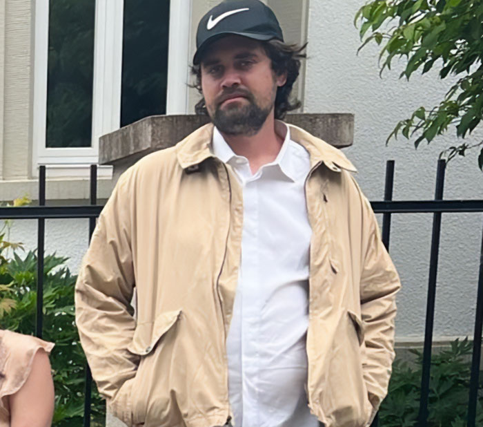 Man wearing beige jacket and black Nike cap standing outdoors near a fence during a casual gathering.