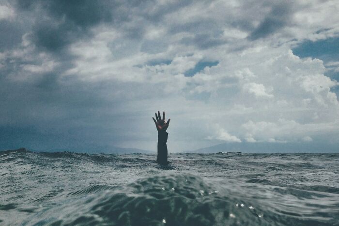 Hand reaching out of turbulent ocean waves under a cloudy sky, illustrating surprising life facts that might make you do a double take.