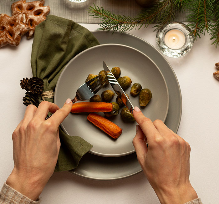 Hands cutting roasted carrots and Brussels sprouts on a plate, illustrating no vegan options Christmas Eve dinner setting.
