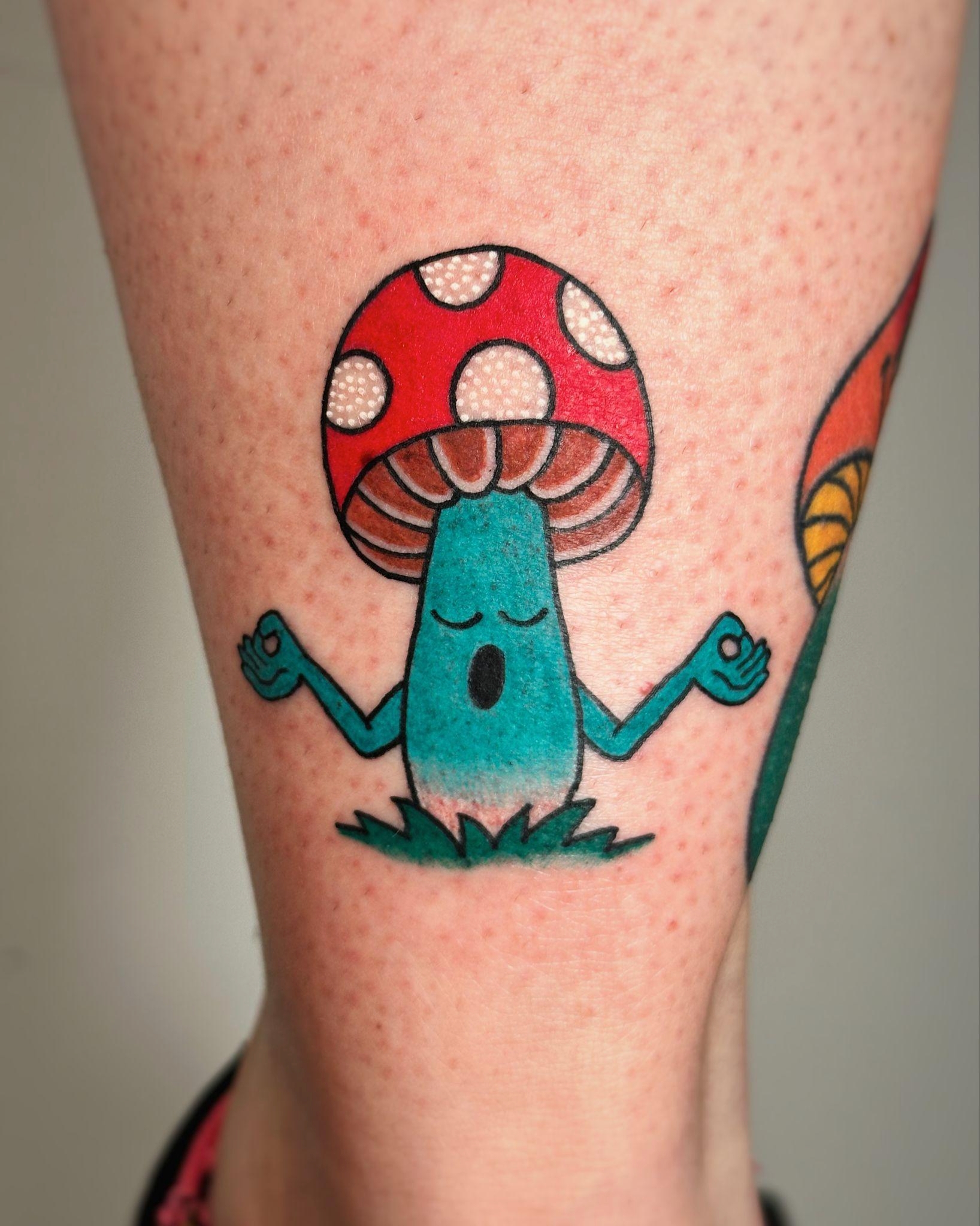 Colorful psychedelic tattoo of a meditating mushroom with a red spotted cap on a person's leg.