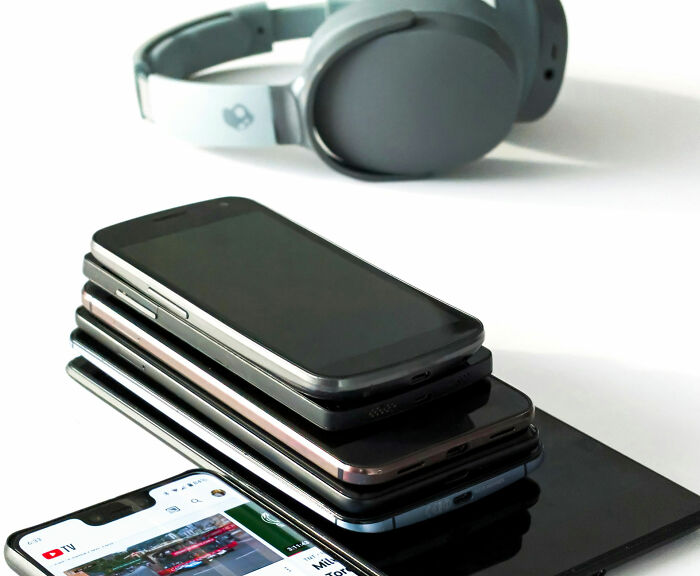 Stack of various smartphones and a tablet with headphones in the background, unusual personal belongings at lost and found.