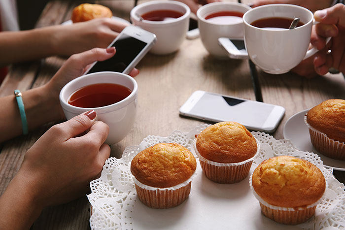 People holding cups of tea at an office potluck with store bought desserts and smartphones on the wooden table.