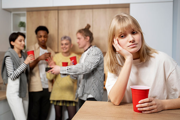 A young woman feeling left out at an office potluck, holding a red cup while others socialize in the background.
